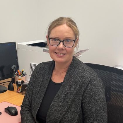 Dr Sanet is sat at her desk wearing a grey blazer and glasses with her hair tied back. she is smiling